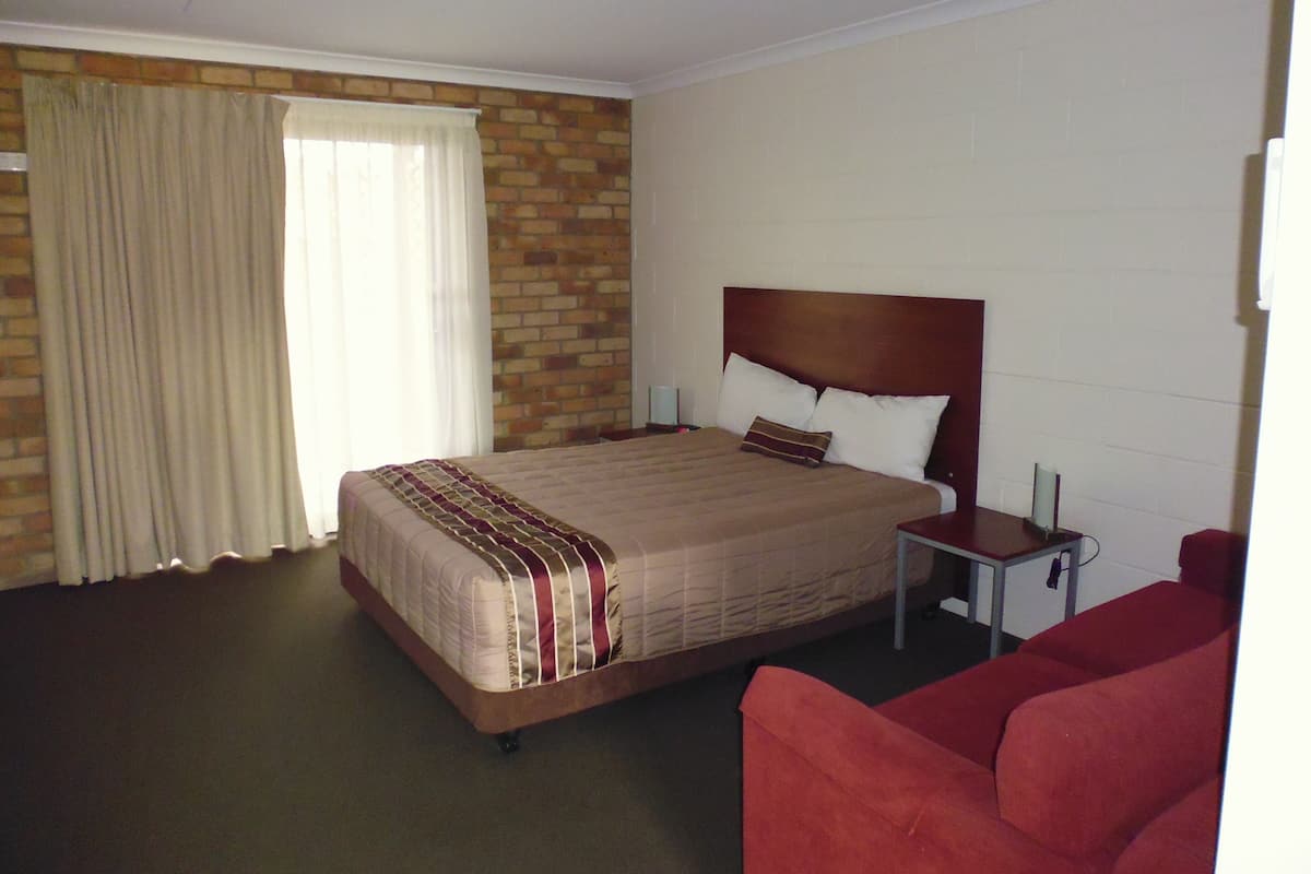 A hotel room with a large bed, a nightstand, a red armchair, and a brick wall.