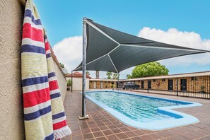 Una piscina con zona sombreada, terraza con baldosas y un edificio con valla al fondo.