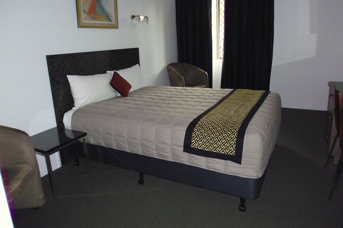 A hotel room with a bed, a chair, a nightstand, a painting on the wall, and a window with curtains.