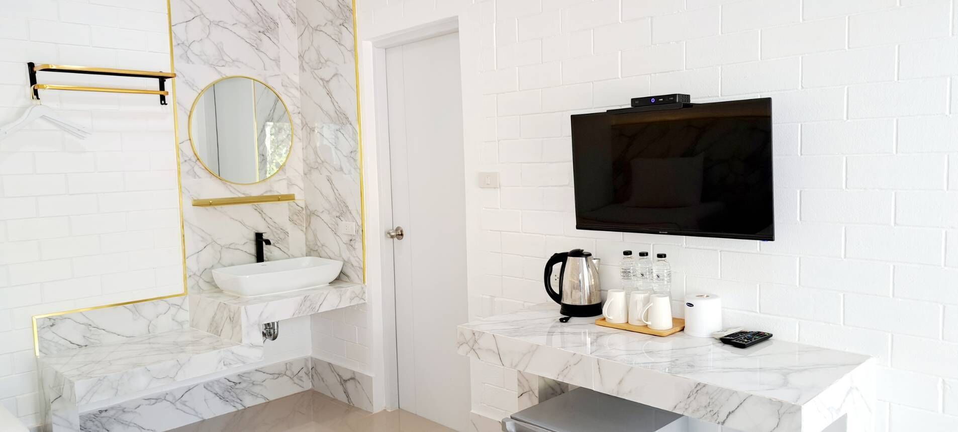 A bathroom with a white marble sink, a gold-framed mirror, a black TV mounted on the wall, and a coffee maker on the countertop.