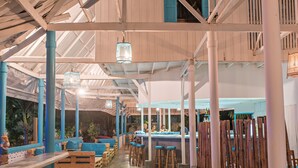 Un restaurant avec de hauts plafonds, des poutres en bois et des touches de bleu. On y trouve des tables et des tabourets en bois, ainsi qu’un coin bar avec des tabourets.