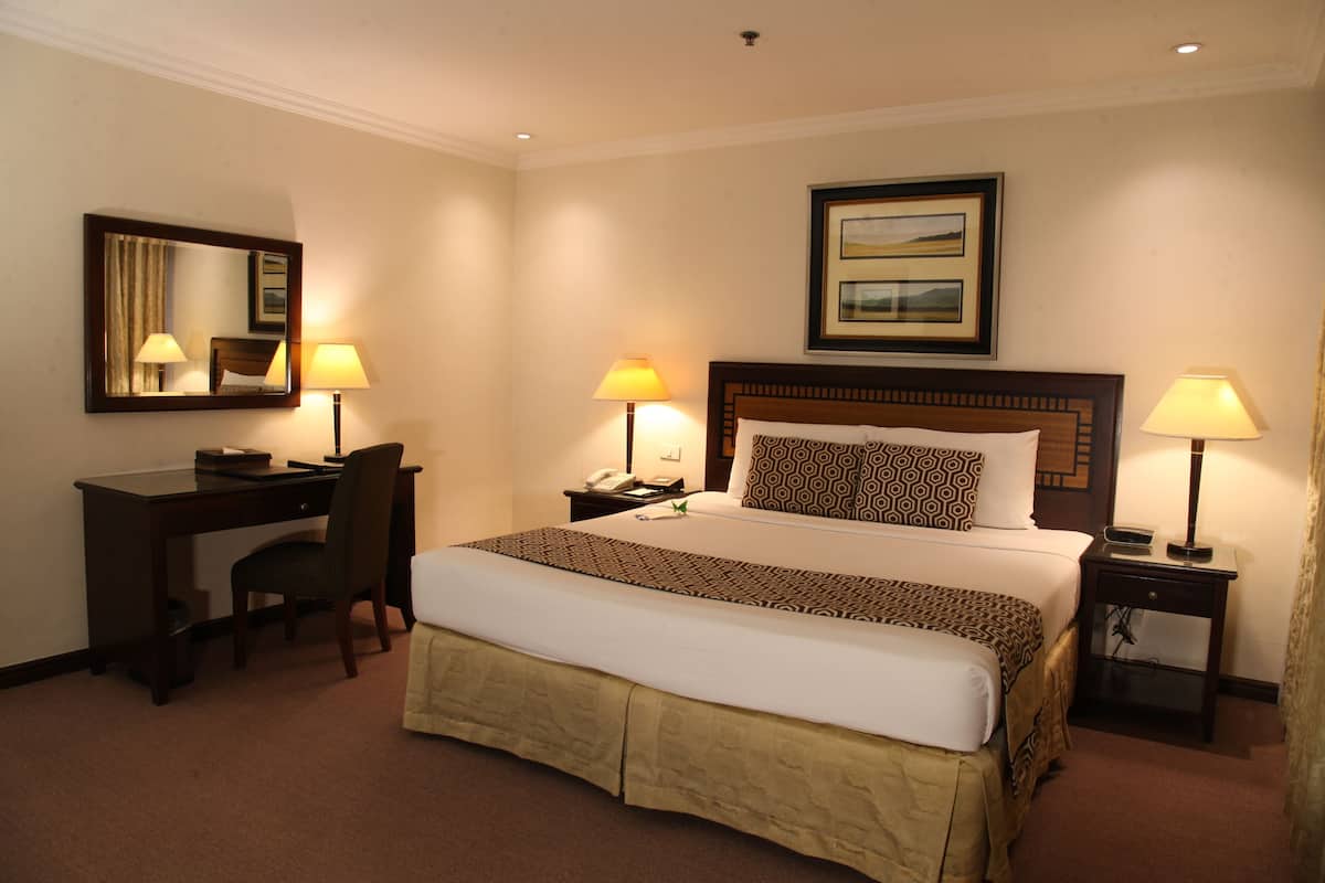 A hotel room with a large bed, two bedside tables with lamps, a desk with a chair, a mirror, and a framed picture on the wall.