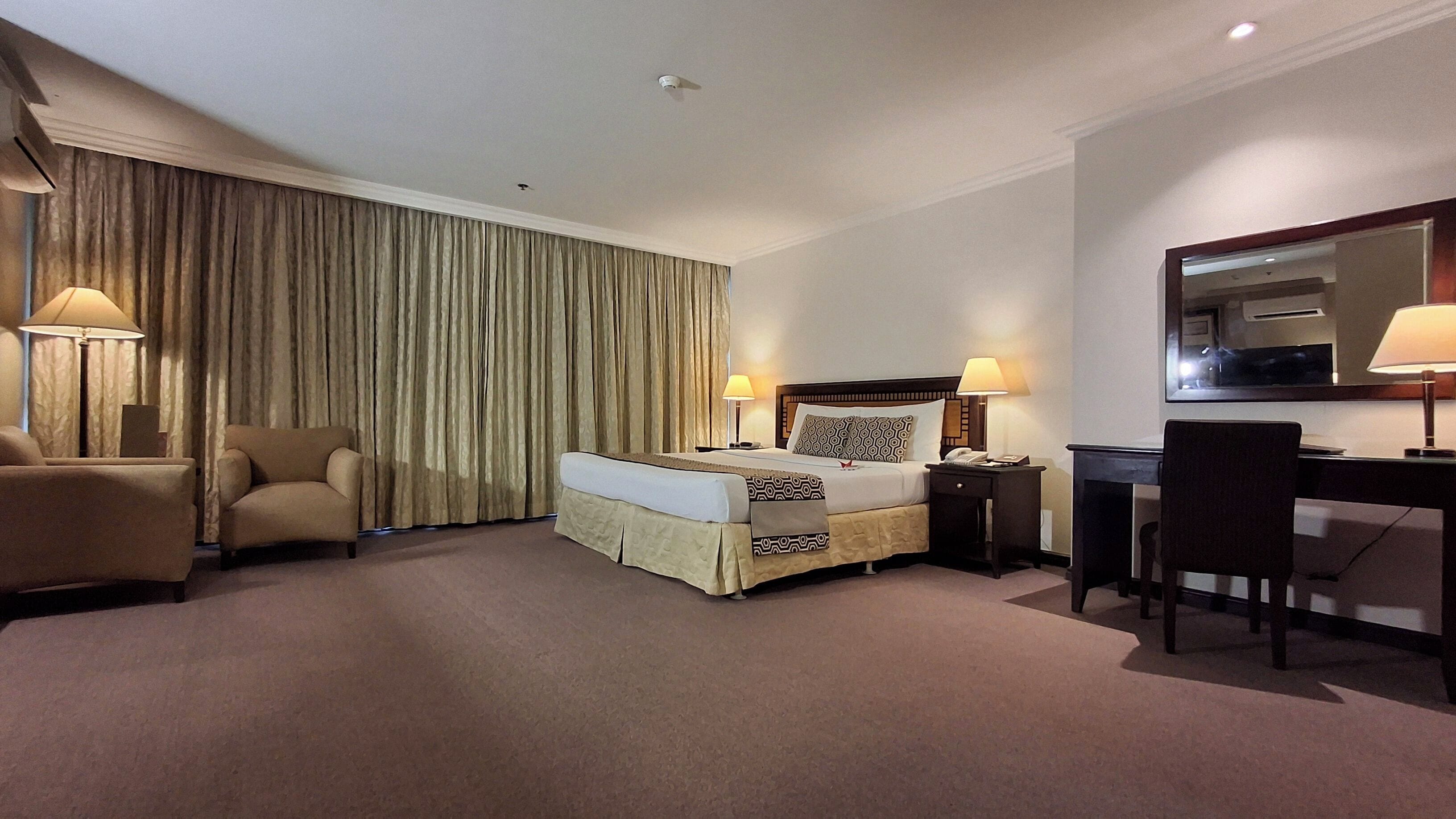 Quarto de hotel com cama, duas luminárias de cabeceira, escrivaninha com cadeira, espelho grande e cortina.