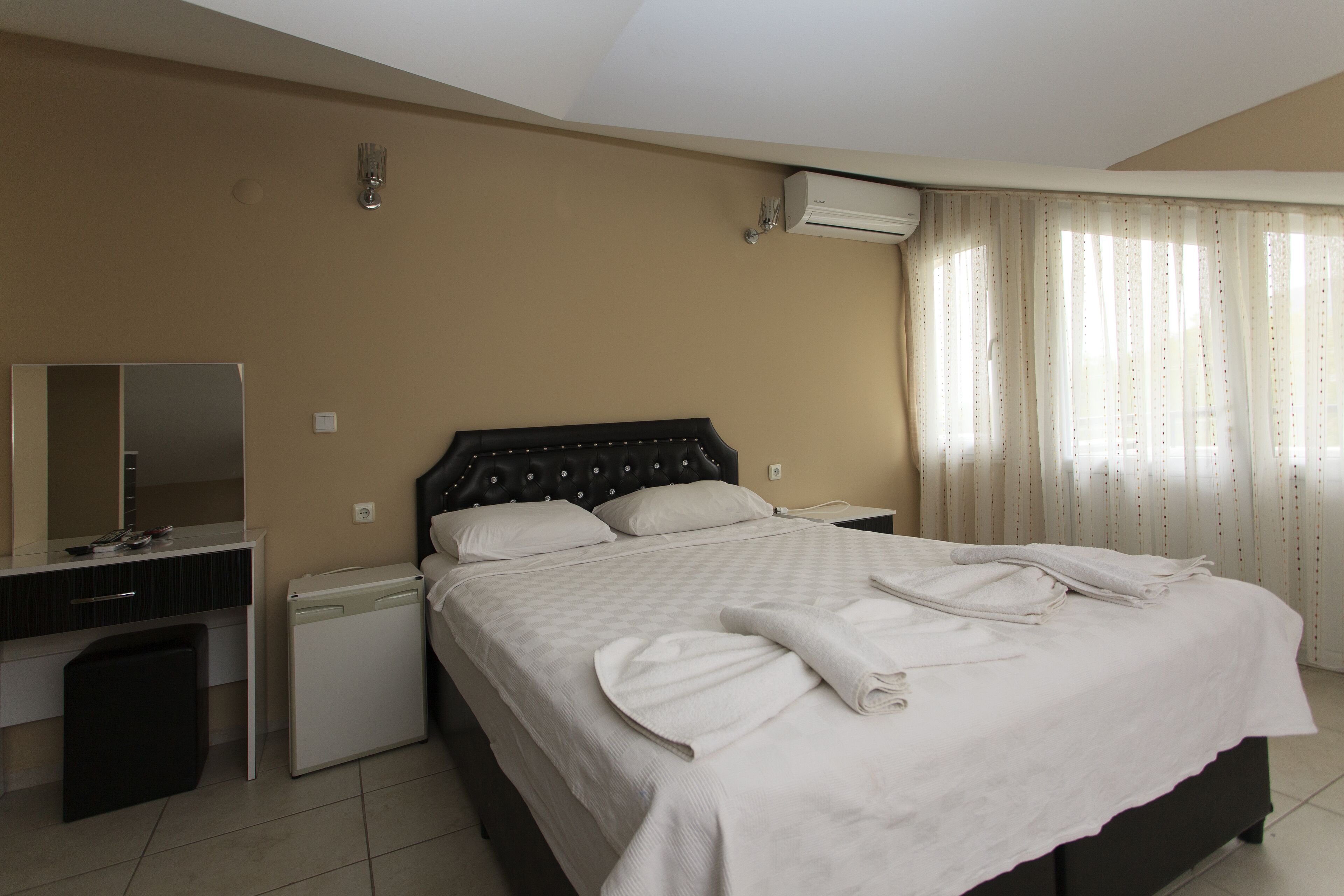 A hotel room with a bed, a desk, a small refrigerator, an air conditioning unit, and curtains.