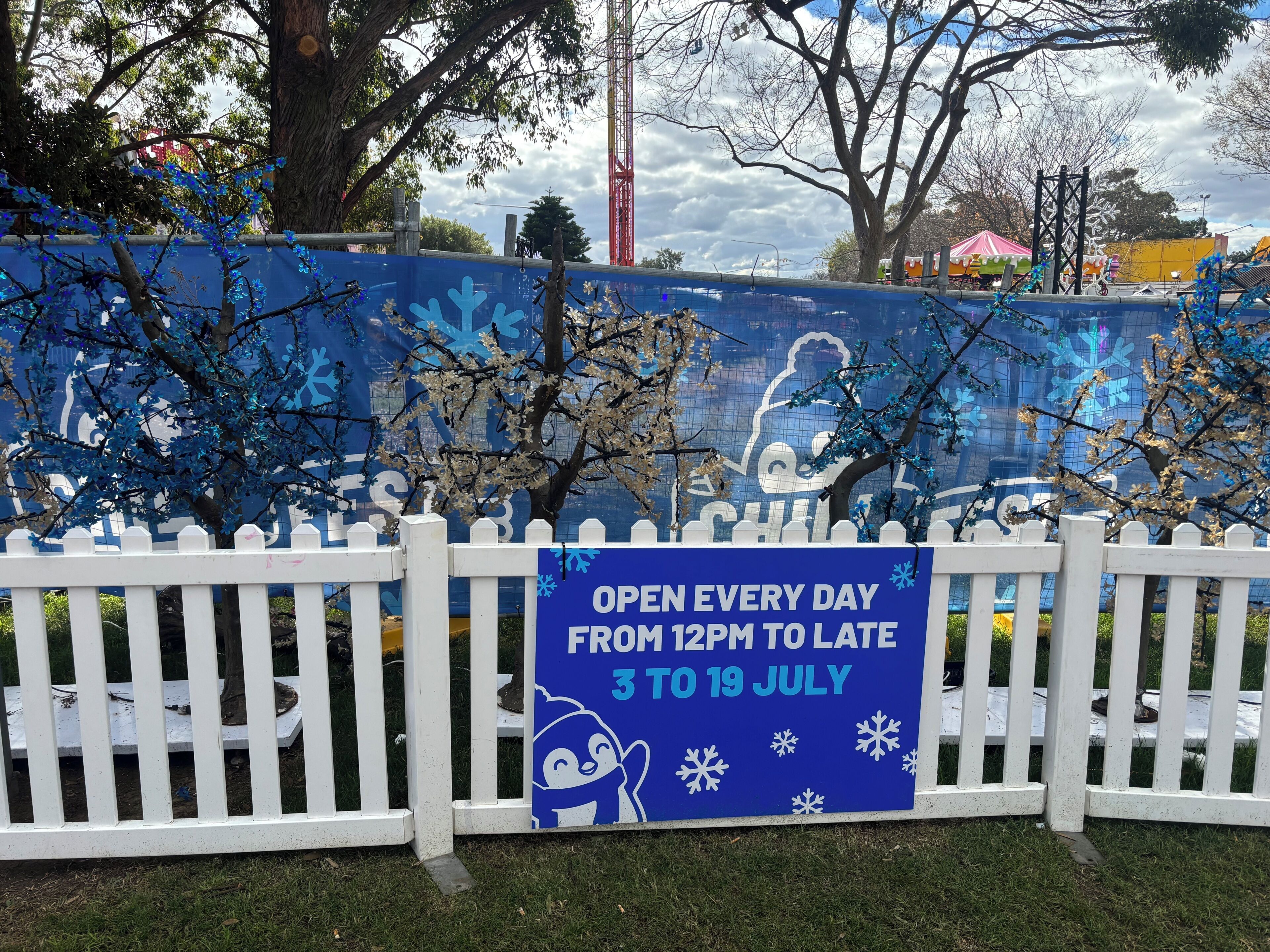 A blue sign with white text stating the opening hours and dates, placed in front of a white picket fence.