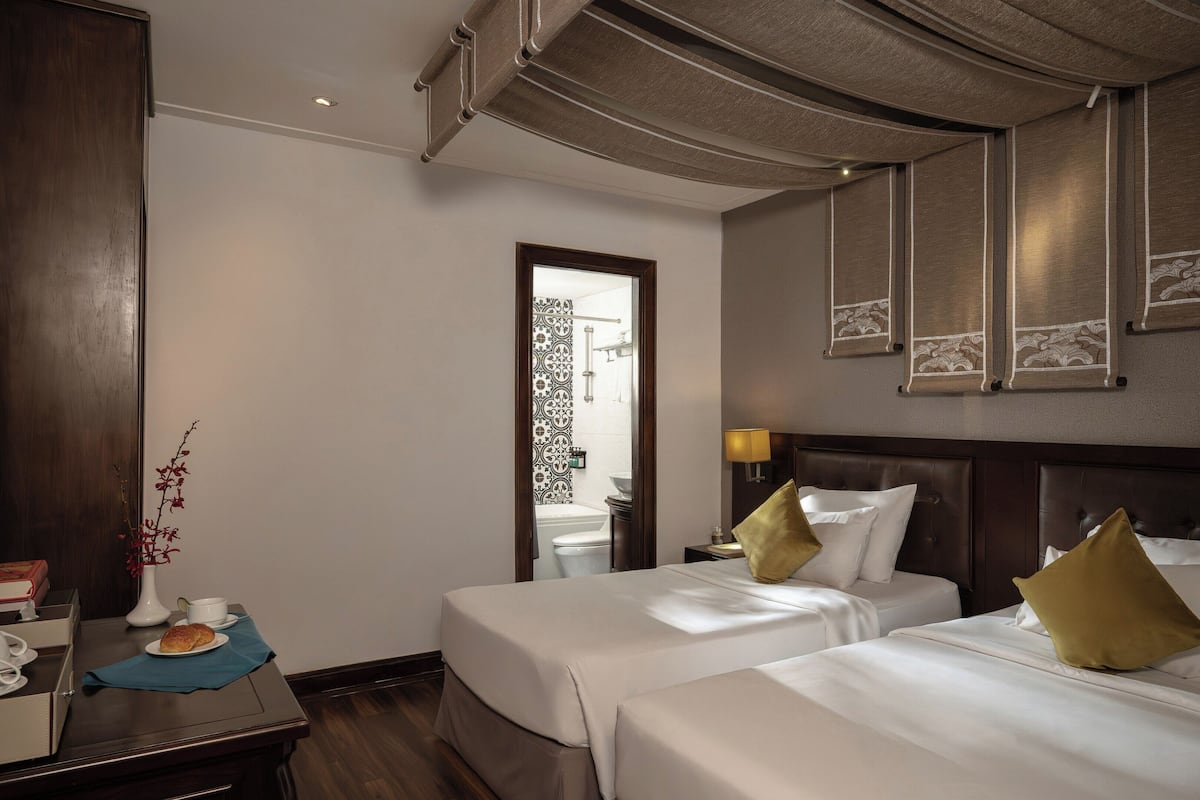 Une chambre d’hôtel avec deux lits, une armoire en bois, un bureau avec un vase et une tasse, et vue sur la salle de bain.
