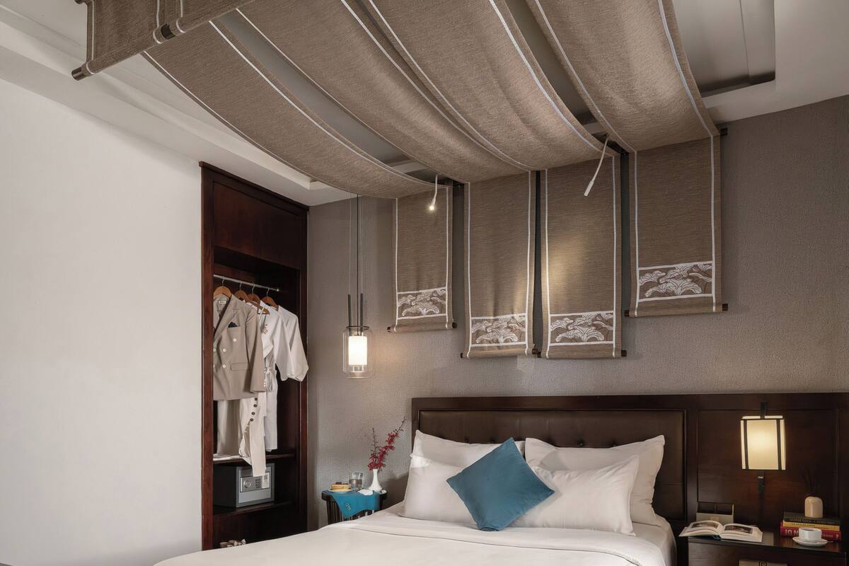 Une chambre d’hôtel avec un grand lit, un plancher en bois, une armoire, une table de chevet avec une lampe et une petite table de nuit avec des livres et une tasse.