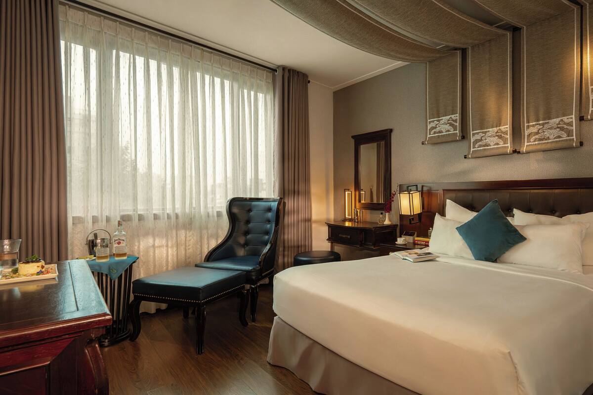 Une chambre d’hôtel avec un grand lit, un bureau avec une chaise, une coiffeuse avec un miroir et une fenêtre avec des rideaux.