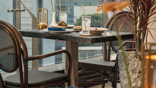 A dining area with a table set for two, chairs, a vase with a plant, and a city view in the background.