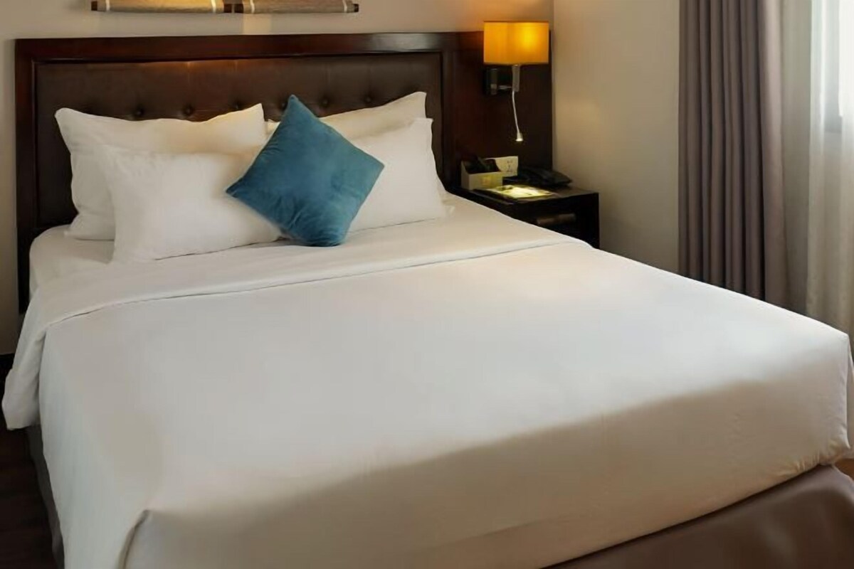 A neatly made bed with white and blue pillows, a wooden headboard, a bedside lamp, and a nightstand with a phone and a small table.