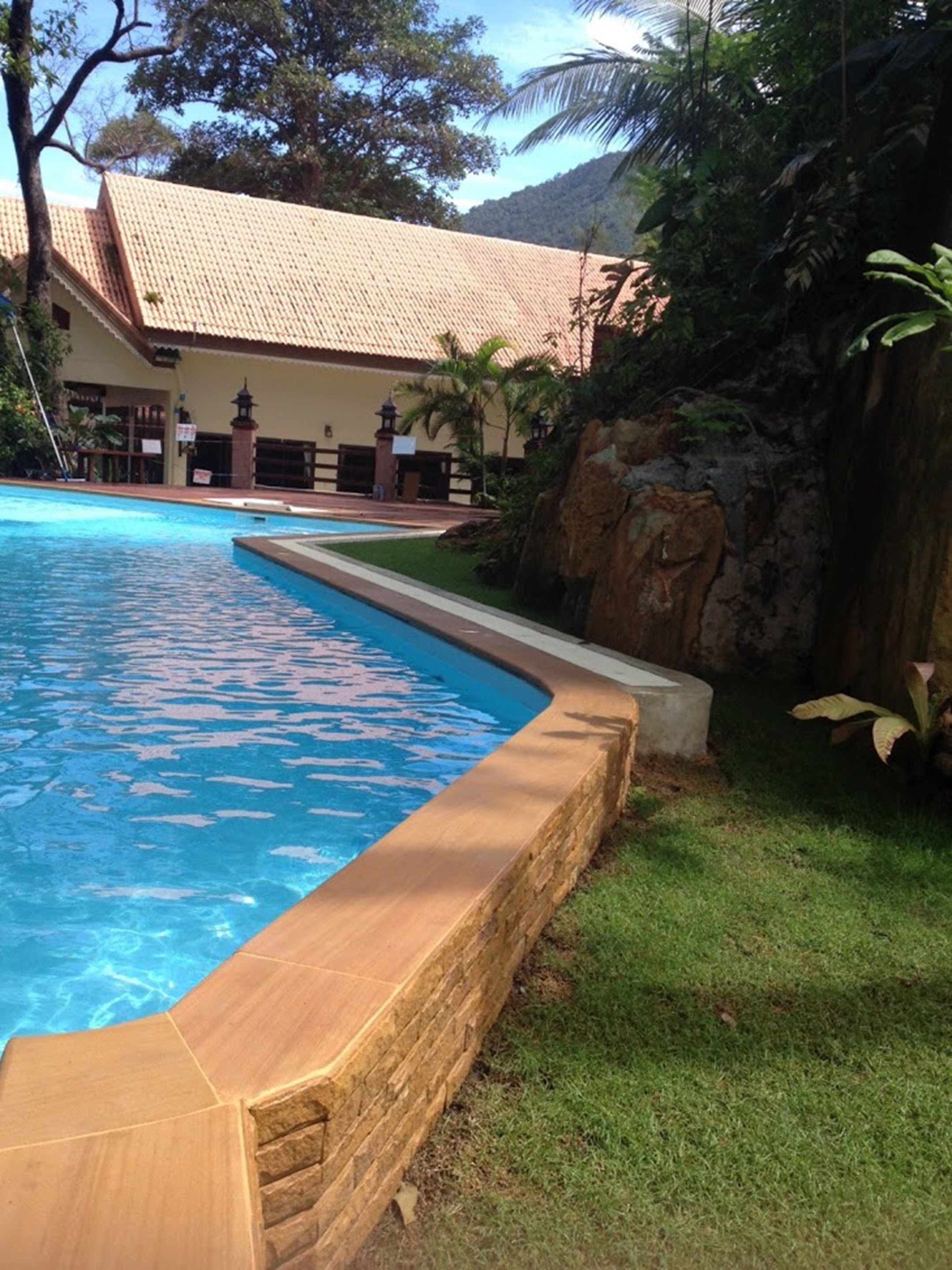A swimming pool with a tiled deck, surrounded by a grassy area and a building with a tiled roof.