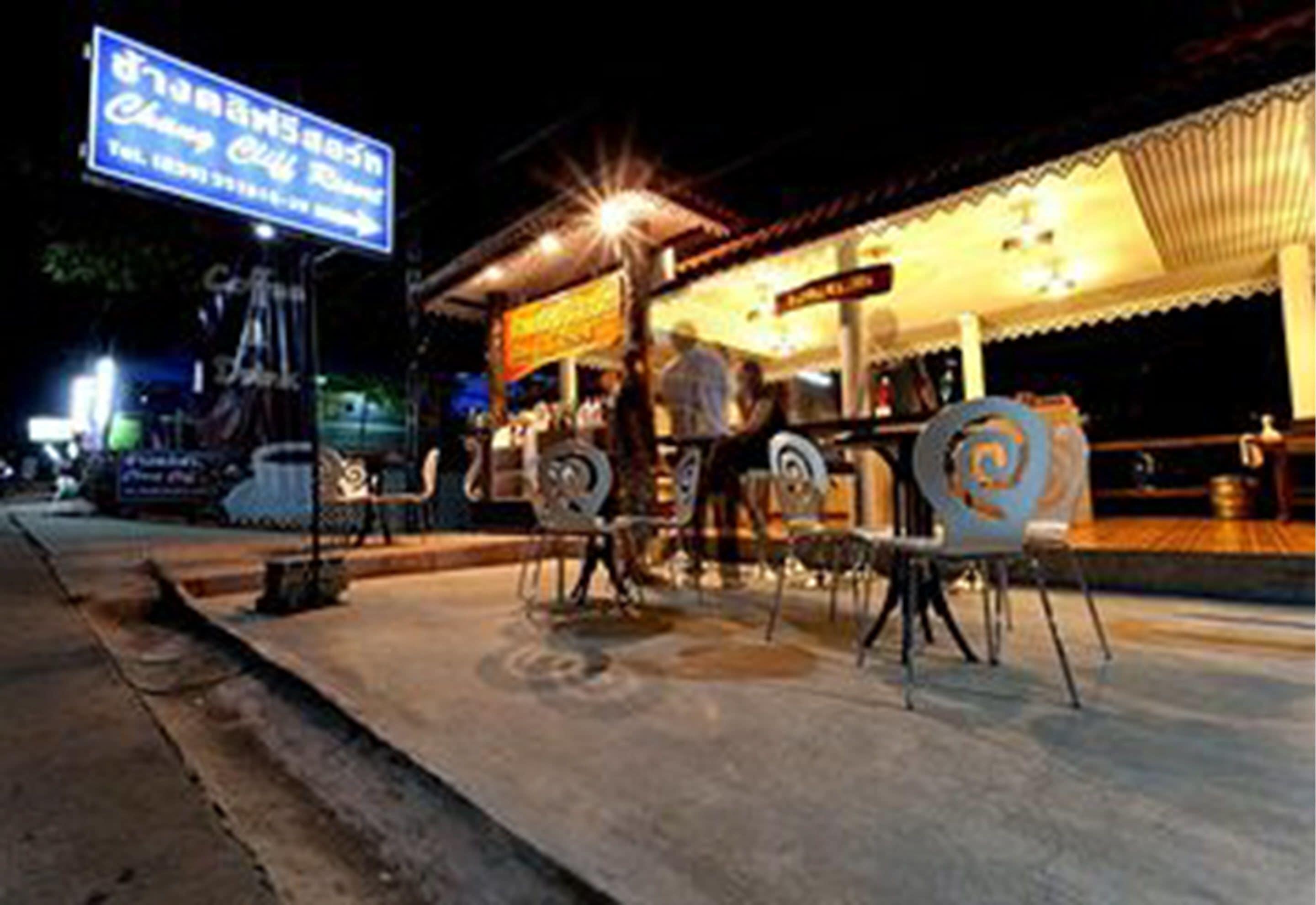 An outdoor seating area with metal chairs and tables, a signboard, and a building with a tiled roof in the background.