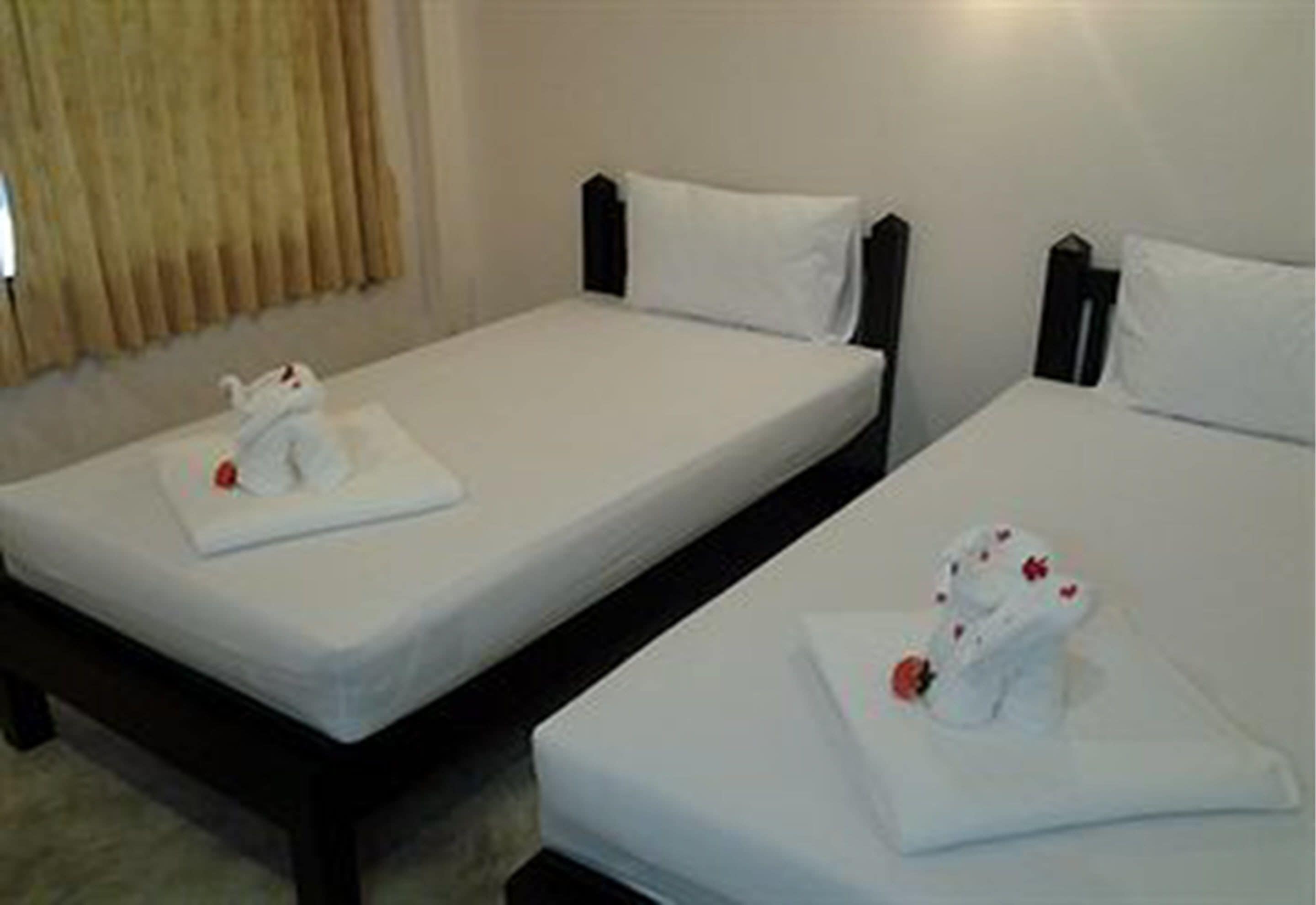 Two single beds with white linens and headboards, a window with beige curtains, and a small table with a white cloth and a red object.