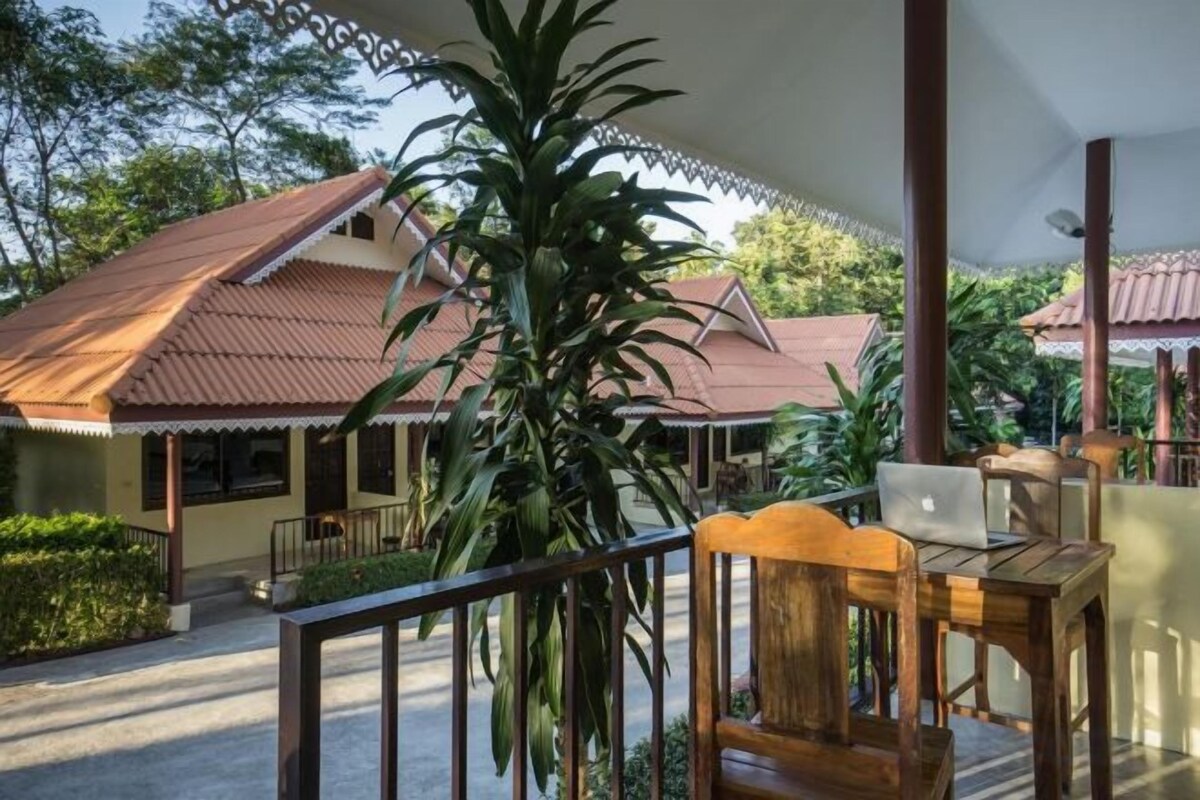 A covered outdoor area with a wooden table and chairs, a laptop, and a large plant.