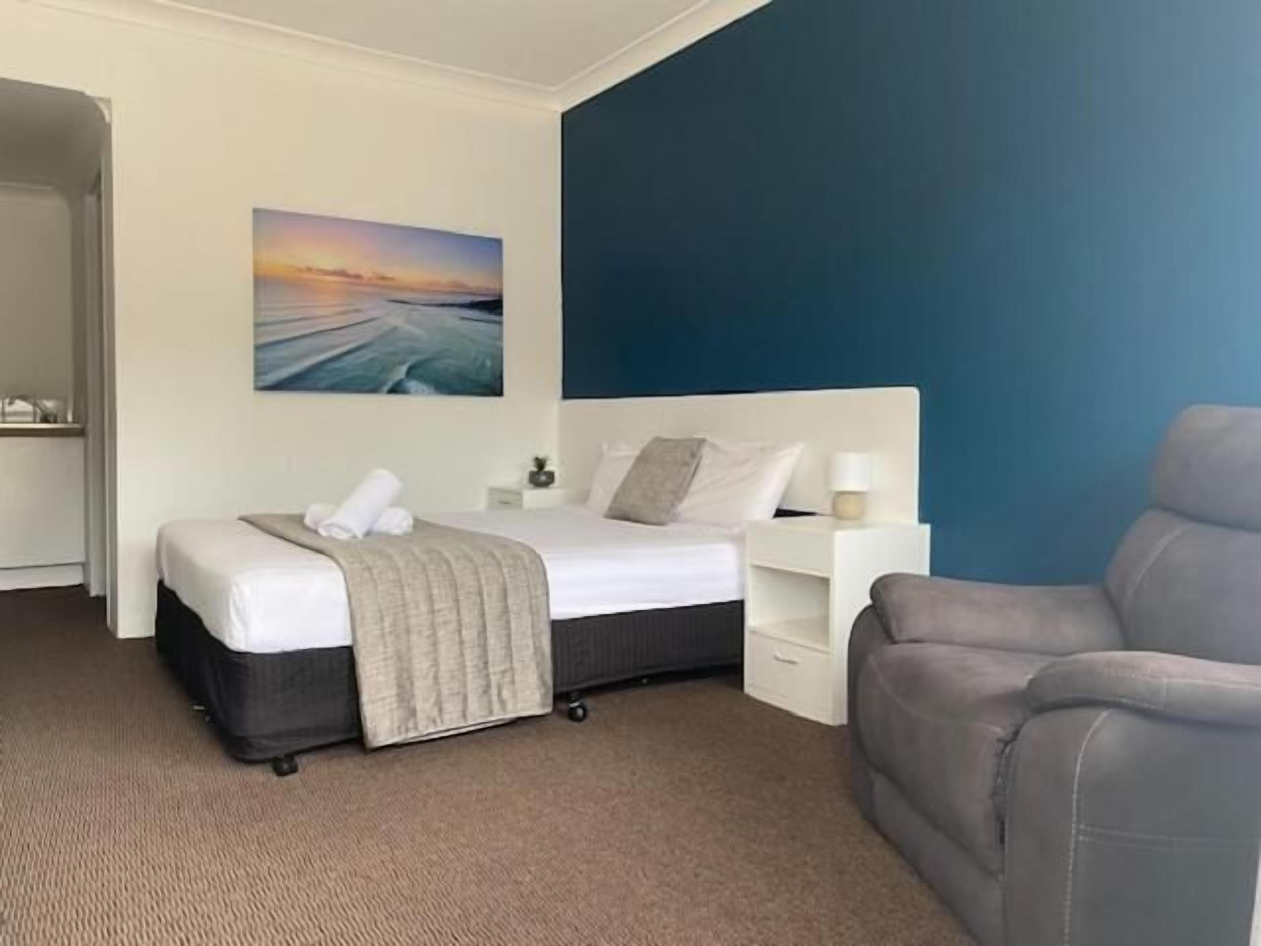 A hotel room with a bed, a gray armchair, a nightstand, and a painting of a sunset over water.
