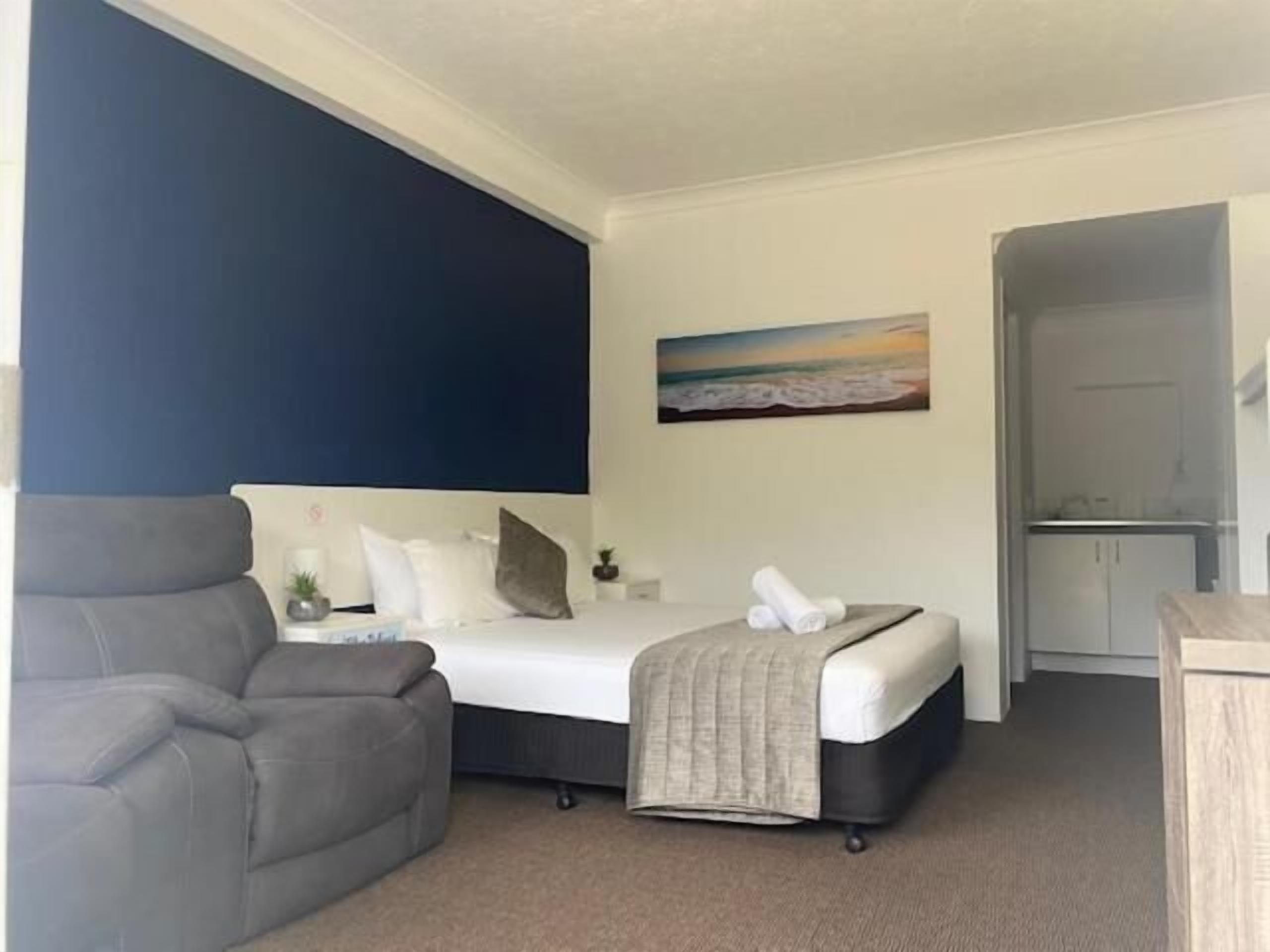 A bedroom with a bed, a grey armchair, a painting of a beach scene, and a door leading to a bathroom.