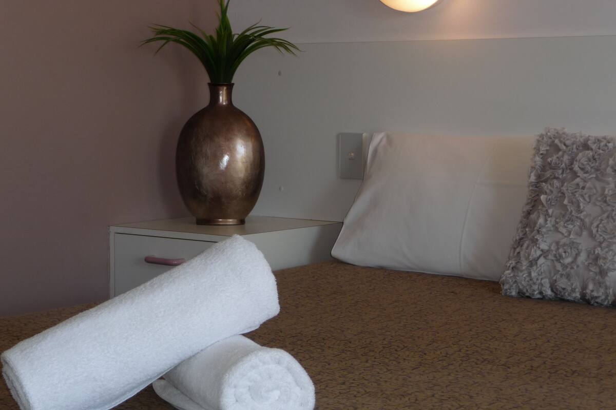 A neatly made bed with white linens, a grey floral pillow, and a brown vase with green plants on a bedside table.