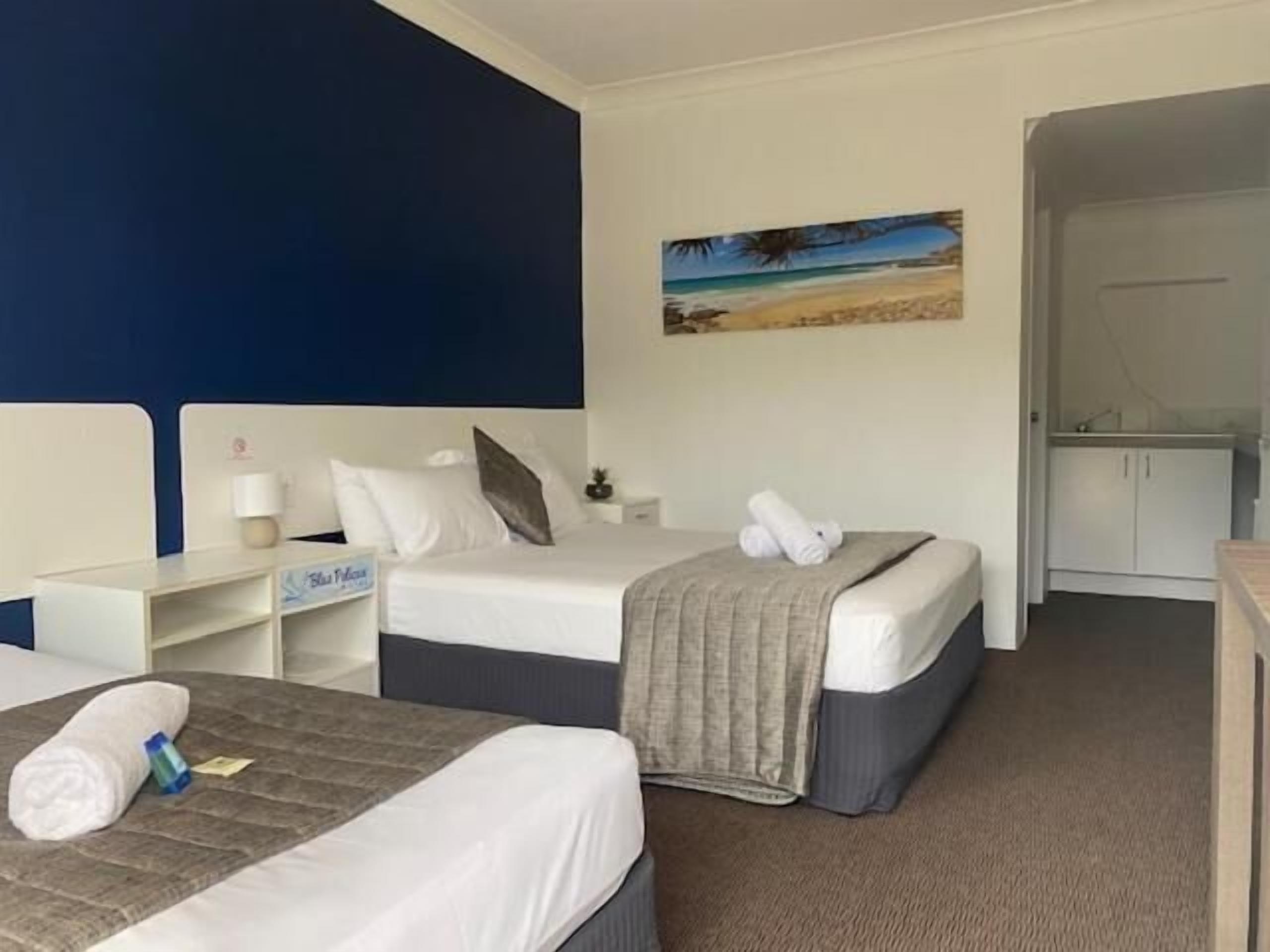 A hotel room with two beds, a painting of a beach, a minibar, and a bathroom visible through an open door.