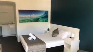 A hotel room with a bed, a nightstand, a desk, and a painting of a coastal scene. - Blue Pelican Motel (Tweed Heads)