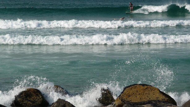 A beach with waves, a person surfing, and rocks in the foreground. - Blue Pelican Motel (Tweed Heads)