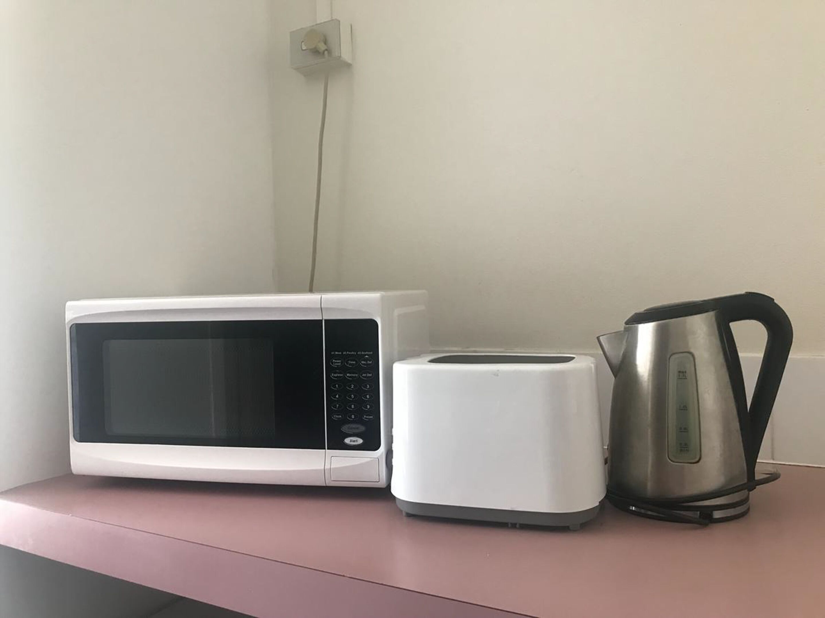 Un microondas, un tostador y una tetera sobre una encimera rosa.