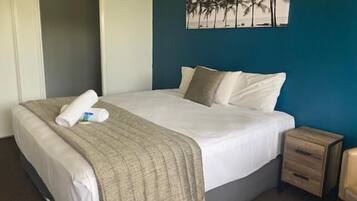 A neatly made bed with a beige blanket and a rolled towel, a small wooden nightstand with drawers, and a wall-mounted painting of palm trees.