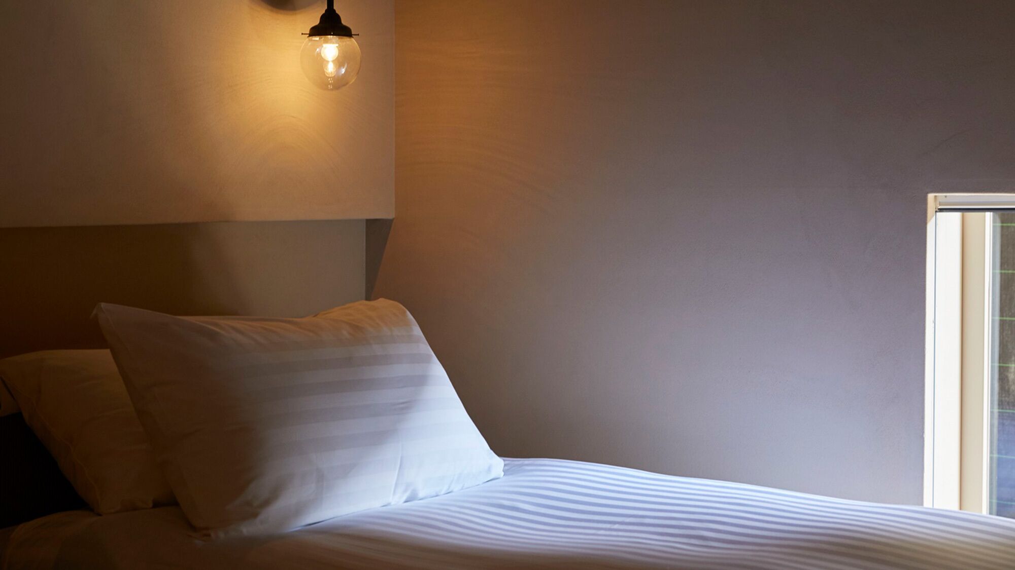 Ein Bett mit weißer Bettwäsche, einem einzelnen Kopfkissen und einer Wandleuchte.