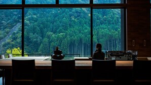 Une pièce dotée de grandes fenêtres donnant sur une forêt dense. On y trouve une table avec des chaises et une machine à café.