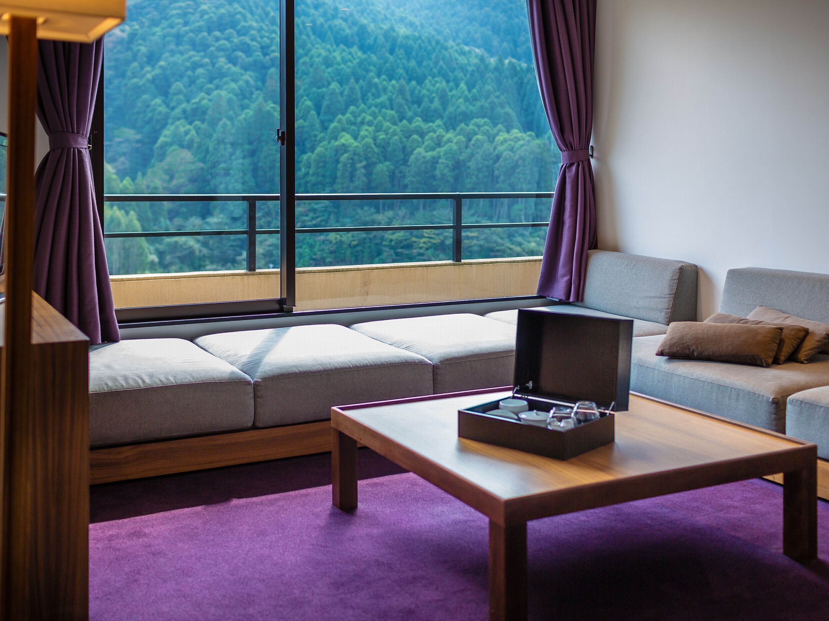 A room with a sofa, a table, and a view of a forest through large windows.