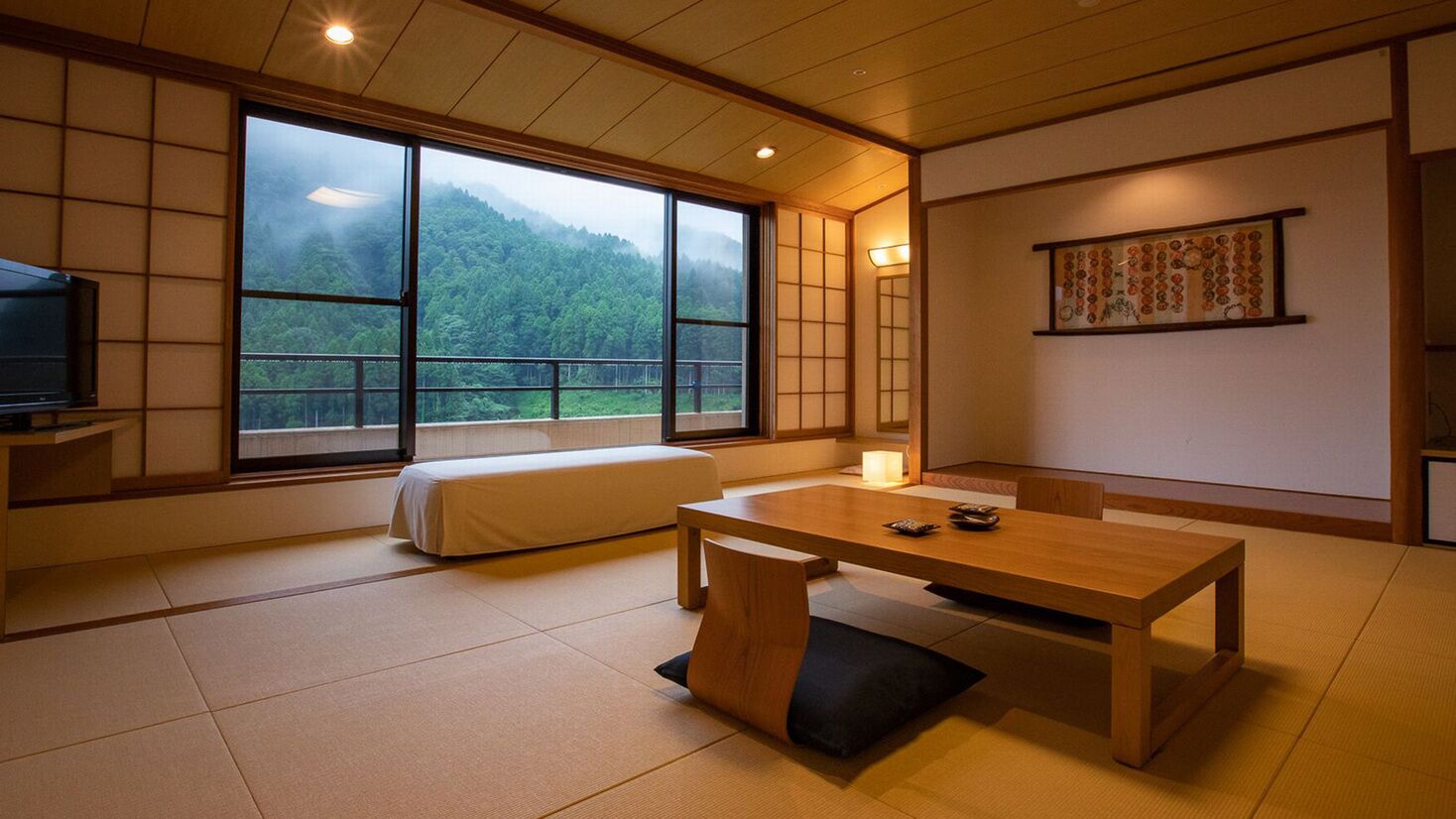 Ein traditioneller japanischer Raum mit Tatami-Boden, einem niedrigen Tisch, einer gepolsterten Sitzbank und einem großen Fenster mit Blick auf einen bewaldeten Berg.