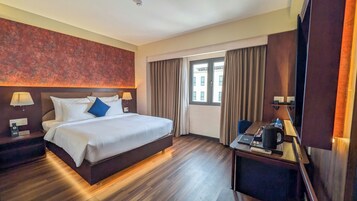A hotel room with a large bed, a desk with a coffee maker, a lamp, and a view of the city through the window.