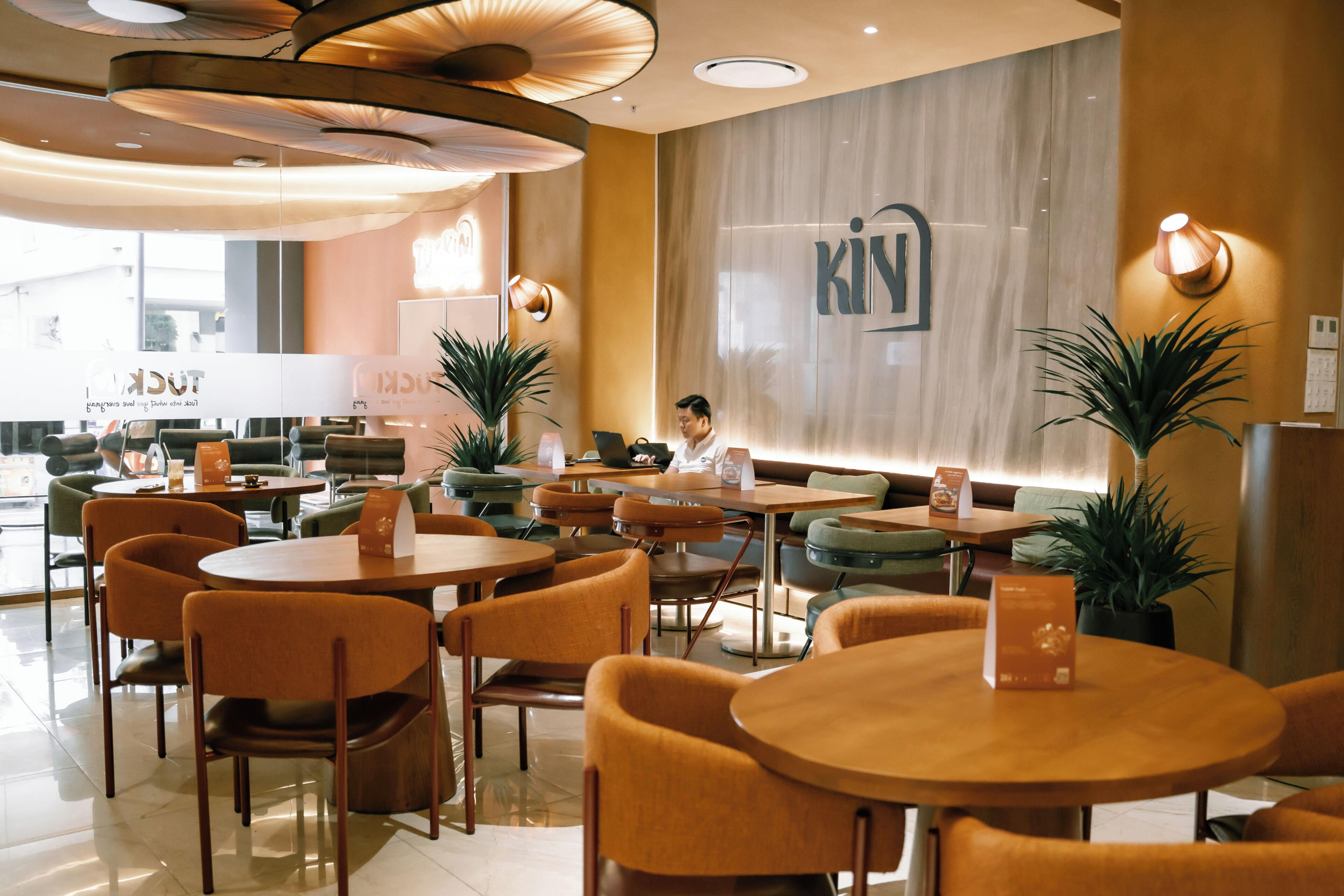 A modern hotel lobby with round tables, orange chairs, and a large 'KIN' sign on the wall.