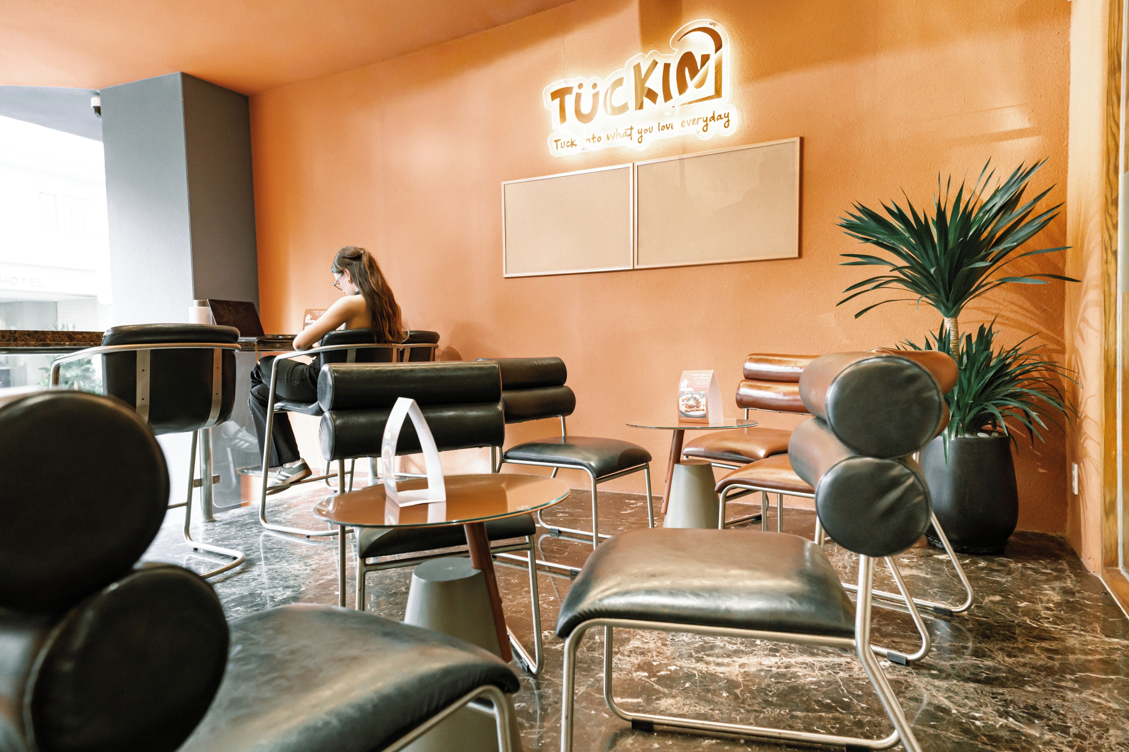 Un interior moderno con sillones de cuero negro, una mesa de centro, una planta en maceta y un letrero que dice 'TUCKIN'.