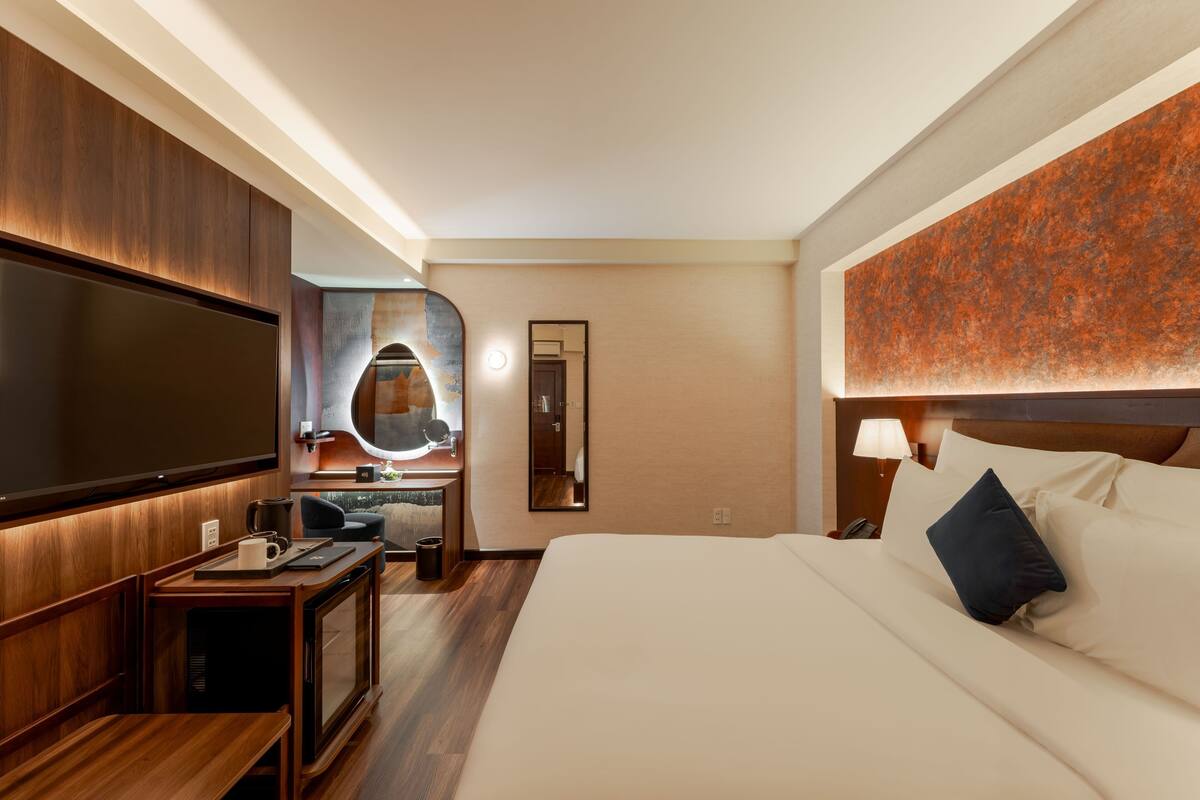 A hotel room with a large bed, a TV, a desk with a chair, a mirror, and a wall-mounted lamp.