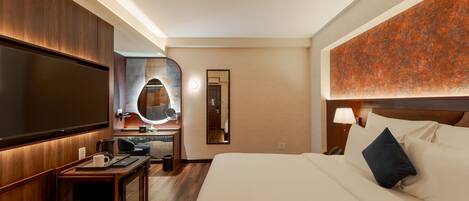 A hotel room with a large bed, a TV, a desk with a chair, a mirror, and a wall-mounted lamp.