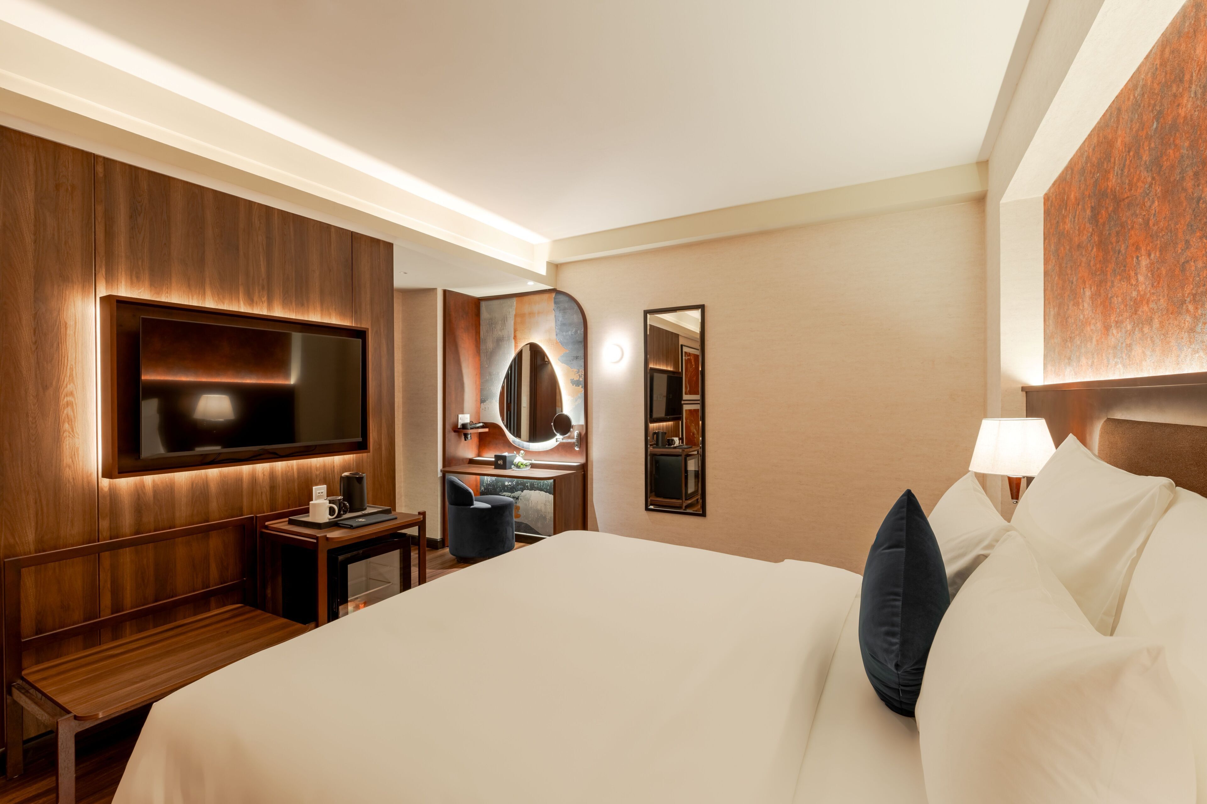 A hotel room with a large bed, a TV mounted on the wall, a desk with a chair, a mirror, and a lamp.