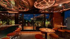 Uma área de bar com mesas redondas e banquetas laranjas, vista para o exterior e design moderno no teto.