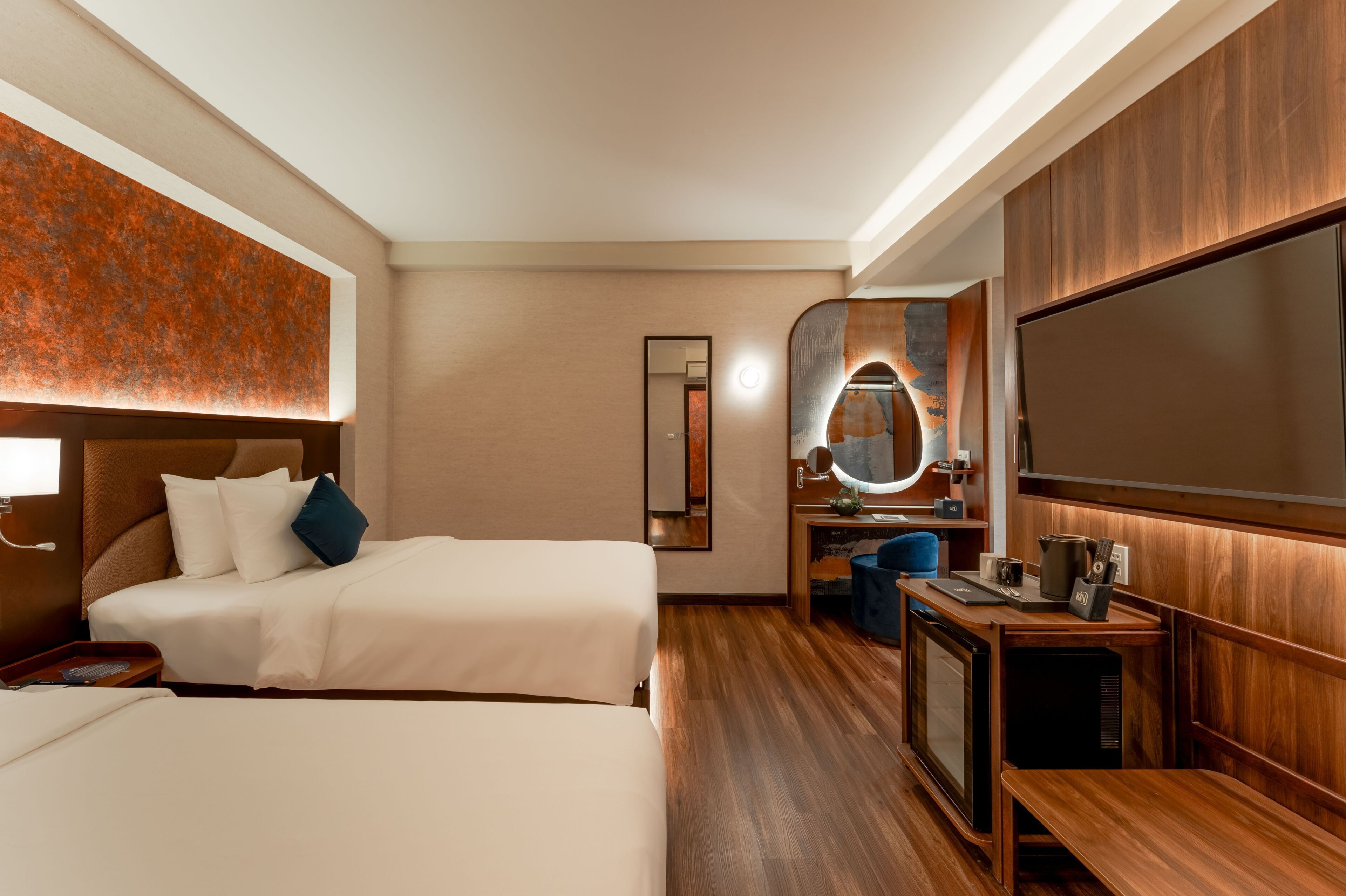 Ein Hotelzimmer mit zwei Betten, einem Fernseher, einem Spiegel und einem kleinen Schreibtisch mit Lampe.