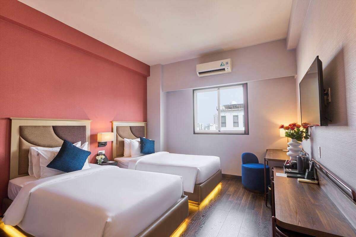 A hotel room with two beds, a desk, a chair, a window with a city view, and an air conditioning unit.