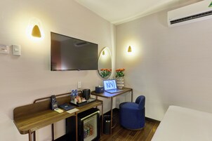 Una camera d'albergo con televisione a schermo piatto, una scrivania con un portatile, un piccolo frigorifero, una sedia blu e un letto.