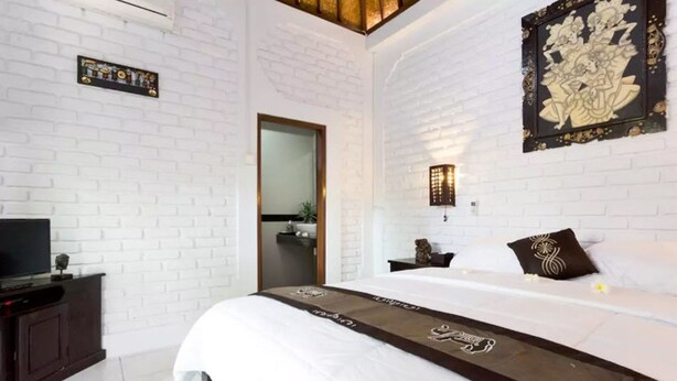 Une chambre d’hôtel moderne avec un mur en briques blanches, un grand lit agrémenté d’un couvre-lit marron, un téléviseur à écran plat posé sur une commode en bois foncé, et un élément décoratif mural au-dessus du lit.