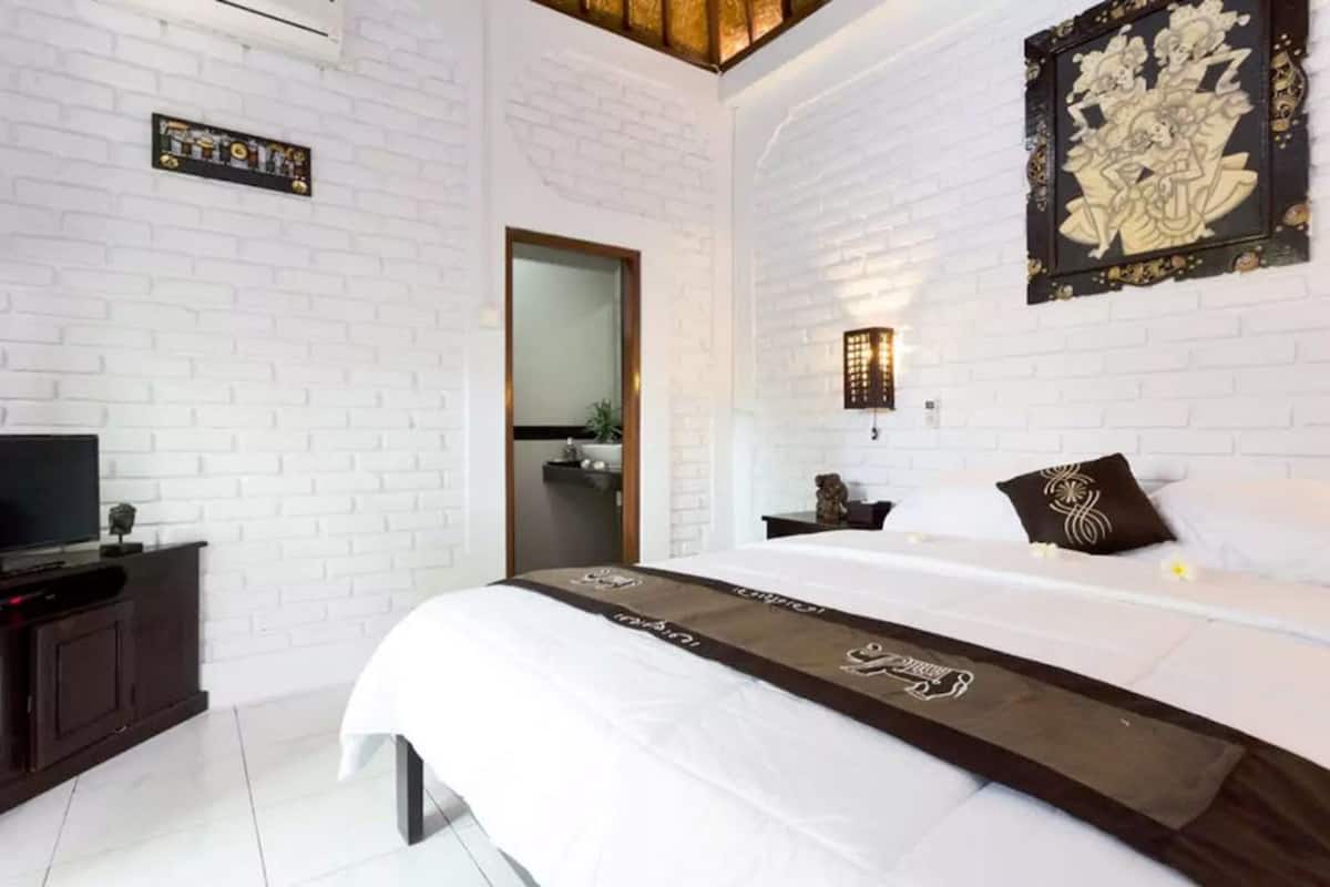 Een moderne hotelkamer met een witte bakstenen muur, een groot bed met een bruine loper, een flatscreen televisie op een donker houten dressoir en een decoratief kunstwerk boven het bed.