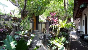 Un étroit sentier bordé d’une végétation tropicale luxuriante et un bâtiment traditionnel doté d’une porte en bois.