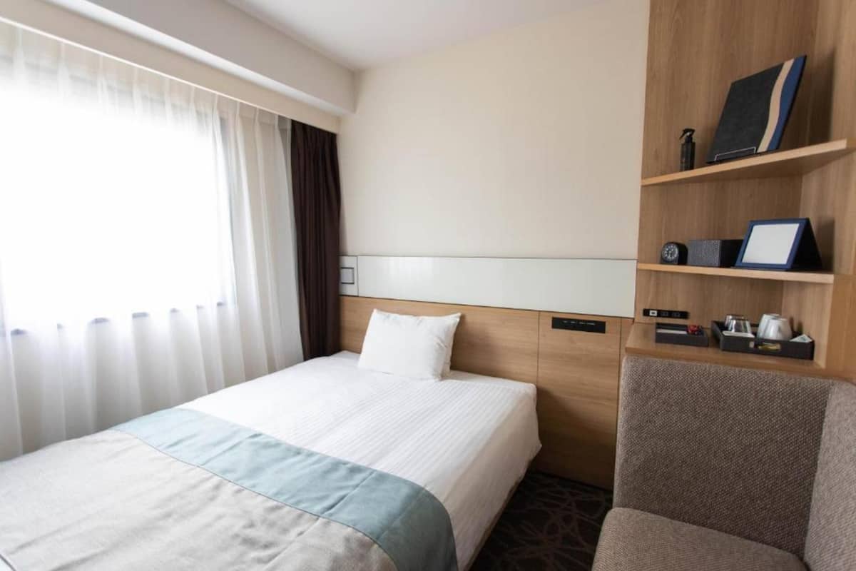 Une chambre d’hôtel avec un lit, une chaise, une étagère avec quelques objets et une fenêtre avec des rideaux.