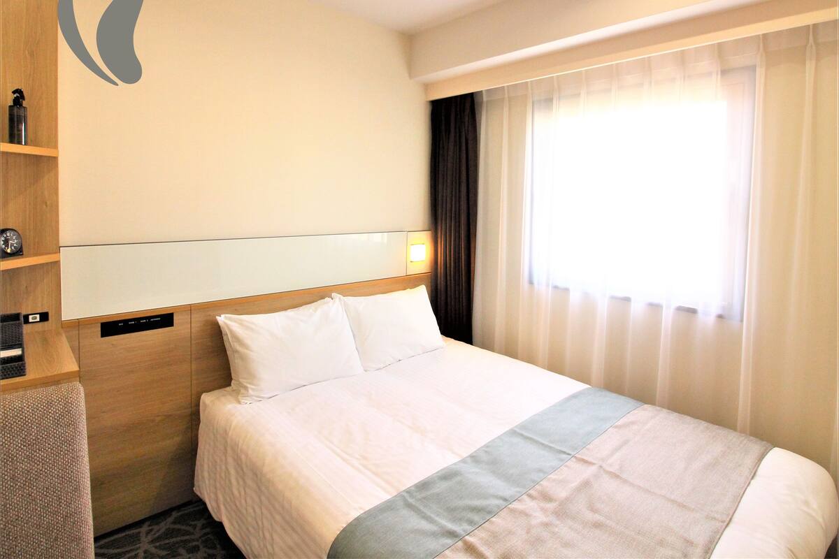 Une chambre d’hôtel avec un lit, une table de chevet avec une horloge, une étagère avec une bouteille et une fenêtre avec des rideaux.