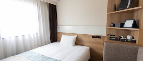 A hotel room with a bed, a chair, a bedside table with a clock, a shelf with a picture frame, and a window with curtains.