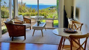 A spacious living area with a large sliding glass door leading to a balcony with a view of the ocean. There are wicker chairs, a small table, and a television on a stand. - Watermark Studio Apartments (Waiheke Island)