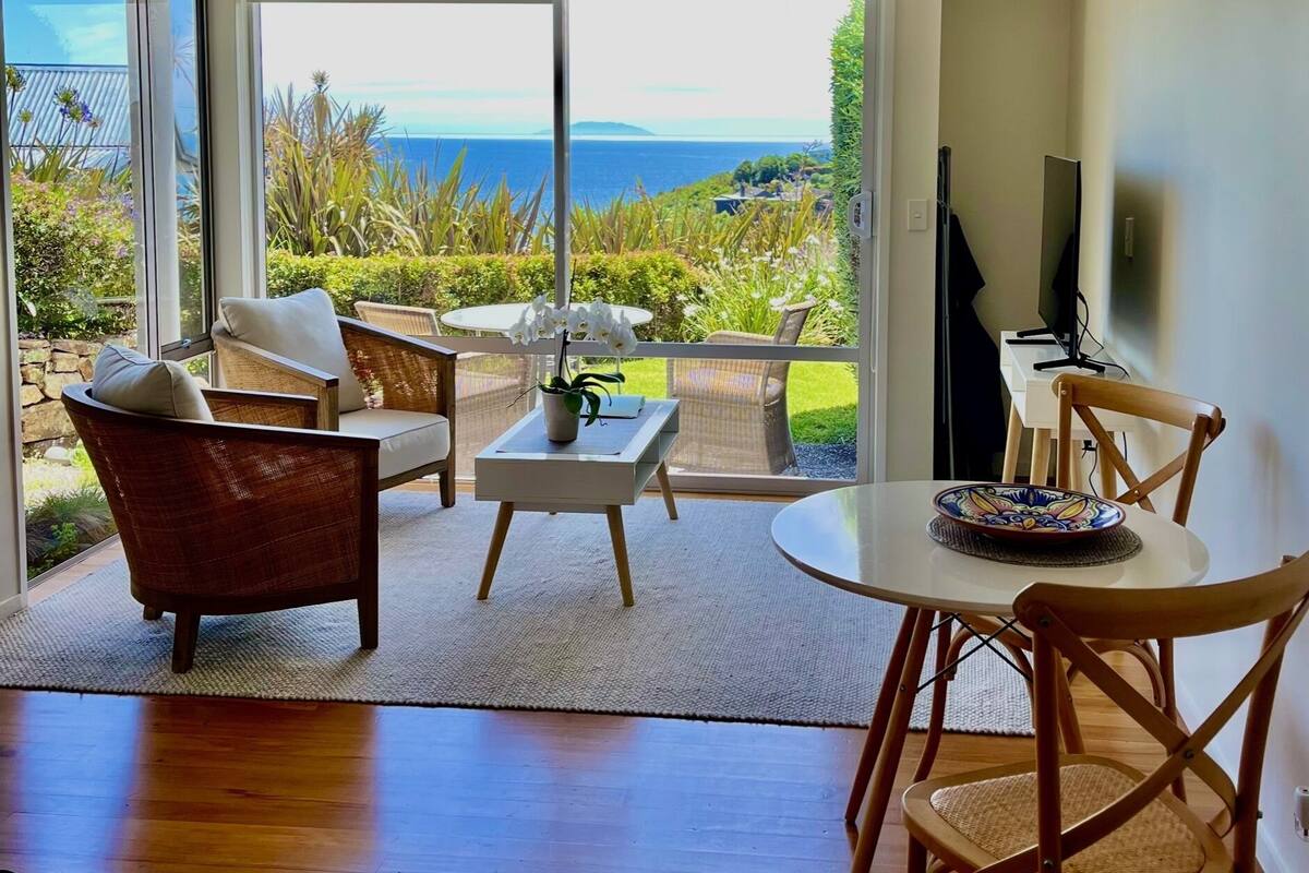 A spacious living area with a large sliding glass door leading to a balcony with a view of the ocean. There are wicker chairs, a small table, and a television on a stand.