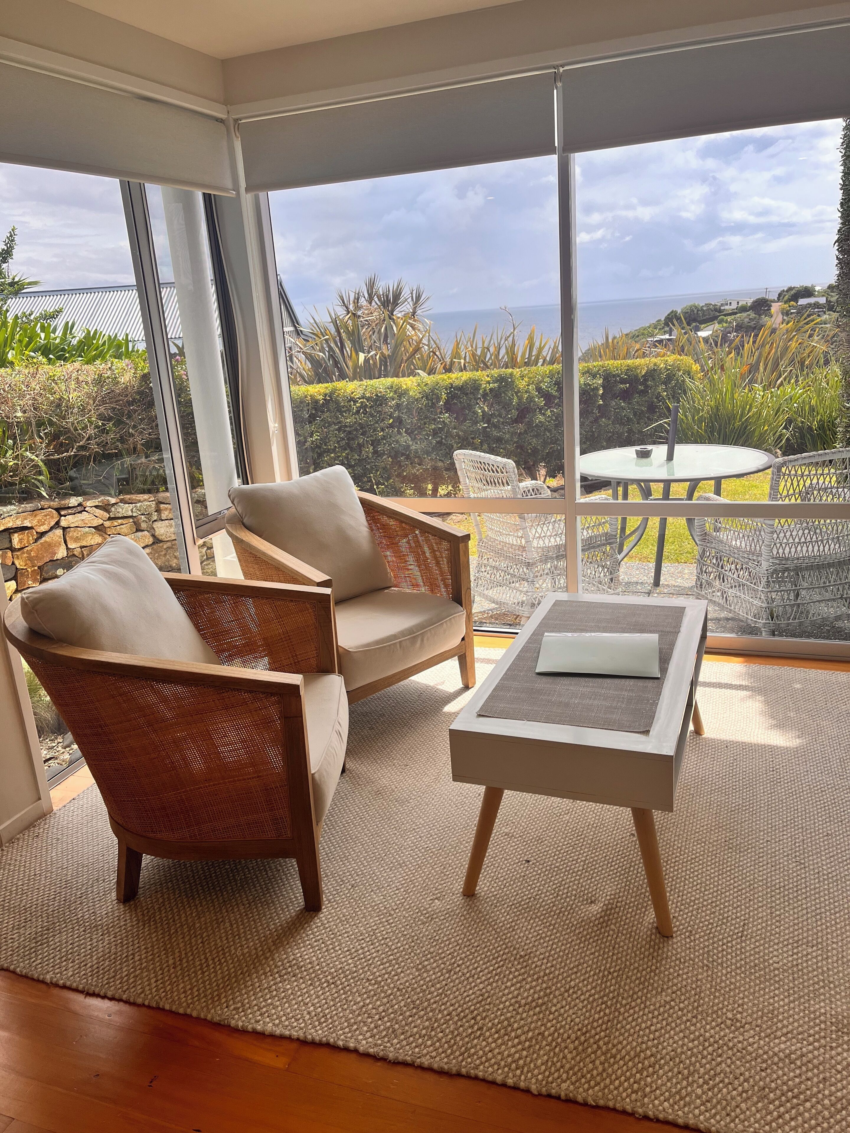 A modern living room with wicker furniture, a glass coffee table, and a view of the ocean and greenery.