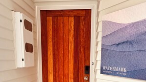 A wooden door with a mailbox and a doormat in front. A sign on the wall reads "Watermark Apartments" with a phone number. - Watermark Studio Apartments (Waiheke Island)